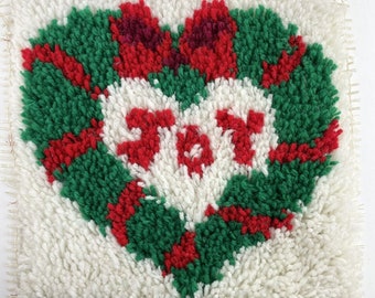 May include: A handmade, square Christmas decoration with a heart-shaped design. The heart is formed by a green wreath with red ribbons and bows. The word "JOY" is stitched in red in the center, set against a white background.