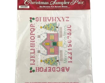 May include: A Bucilla Stamped Cross Stitch Christmas Sampler Pair kit. The design includes a house, Christmas trees, the alphabet, and numbers. The kit is packaged in a clear plastic bag.