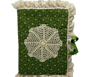 May include: A rectangular fabric-covered journal featuring a green background with a small white and red floral pattern. The cover is decorated with a central cream-colored crocheted doily and is edged with ruffled cream lace and a green ribbon.
