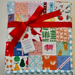 May include: A colourful patchwork quilt tied with a red ribbon. The quilt is made up of squares with animal and fruit patterns. A tag from "Red Bird Quilt Shop" is attached, with text in English.