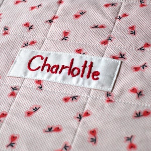 Könnte beinhalten: Eine weiße Stoff-Babydecke mit einem rosa-roten Blumenmuster. Die Decke hat ein weißes Etikett, das mit dem Namen "Charlotte" in roter Farbe bestickt ist.
