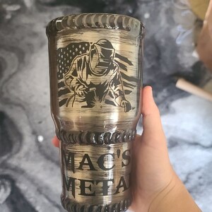 以下が含まれることがあります： 溶接工とアメリカ国旗をあしらったデザインのステンレス製タンブラー。タンブラーには「MAC'S METAL」の文字があり、手で持たれています。