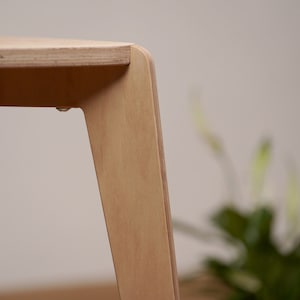 K&ouml;nnte beinhalten: Nahaufnahme eines hellen Holzhockers mit abgerundeten Kanten und einer glatten Oberfl&auml;che. Der Hocker hat drei Beine und ist aus Sperrholz gefertigt.