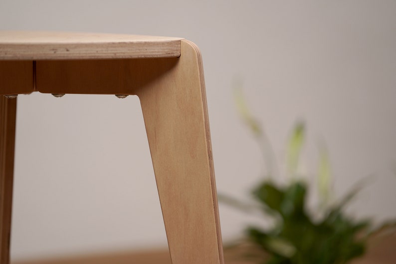 K&ouml;nnte beinhalten: Nahaufnahme eines hellen Holzhockers mit abgerundeten Kanten und einer glatten Oberfl&auml;che. Der Hocker hat drei Beine und ist aus Sperrholz gefertigt.