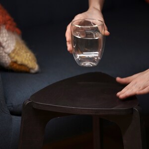 K&ouml;nnte beinhalten: Eine Hand h&auml;lt ein klares Glas mit Wasser &uuml;ber einem dunkelbraunen dreieckigen Tisch.