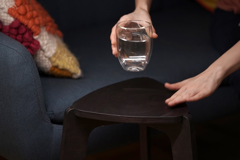 K&ouml;nnte beinhalten: Eine Hand h&auml;lt ein klares Glas mit Wasser &uuml;ber einem dunkelbraunen dreieckigen Tisch.
