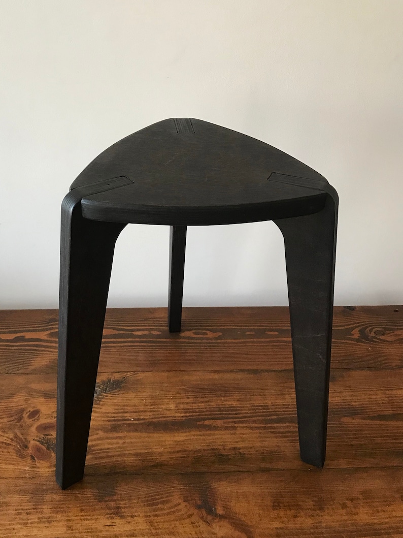 Puede incluir: Un taburete triangular negro con tres patas. El taburete est&aacute; hecho de madera y tiene un acabado liso y pulido.