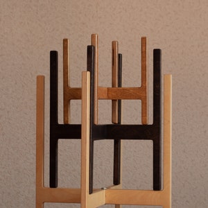 K&ouml;nnte beinhalten: Ein Set aus drei ineinander stapelbaren Pflanzenst&auml;ndern aus Holz mit kreuzf&ouml;rmigem Sockeldesign. Die St&auml;nder bestehen aus Sperrholz mit unterschiedlichen Oberfl&auml;chen, darunter helles Naturholz, eine mittelbraune Beize und eine dunkelbraune Beize.
