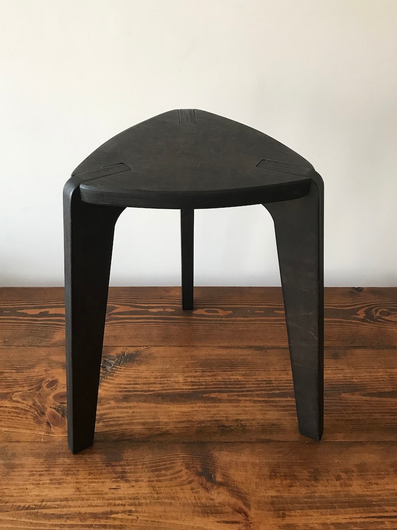Puede incluir: Un taburete triangular negro con tres patas. El taburete est&aacute; hecho de madera y tiene un acabado liso y pulido.