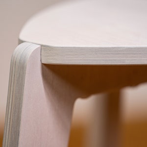 K&ouml;nnte beinhalten: Nahaufnahme eines hellen Holzstuhls mit abgerundeter Oberseite und glatter, wei&szlig;er Oberfl&auml;che. Der Stuhl ist aus Sperrholz gefertigt und hat ein schlichtes, modernes Design.