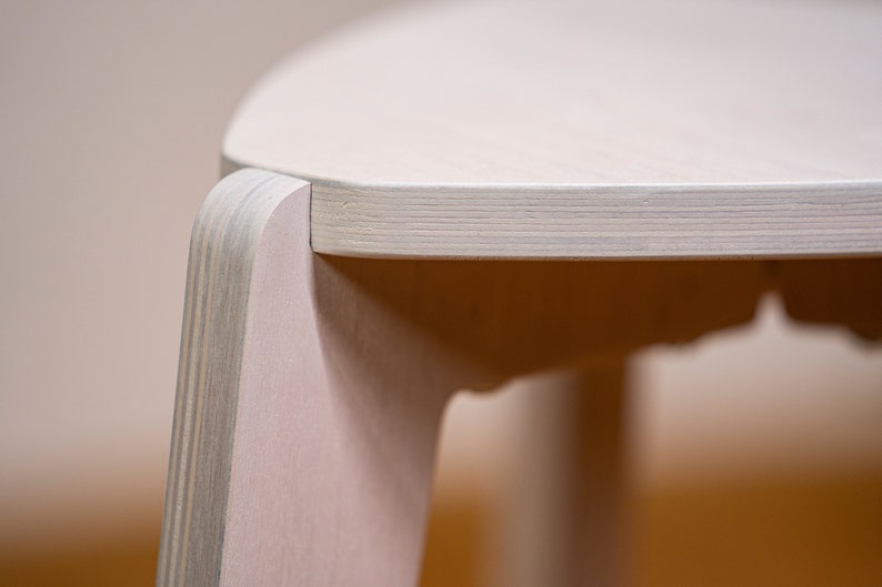 K&ouml;nnte beinhalten: Nahaufnahme eines hellen Holzstuhls mit abgerundeter Oberseite und glatter, wei&szlig;er Oberfl&auml;che. Der Stuhl ist aus Sperrholz gefertigt und hat ein schlichtes, modernes Design.