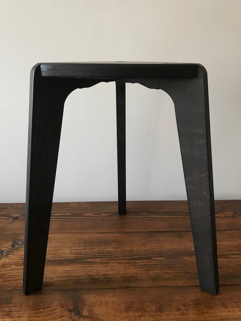 Puede incluir: Un taburete de madera negro con un dise&ntilde;o simple y moderno. El taburete tiene una parte superior rectangular y cuatro patas que est&aacute;n ligeramente inclinadas hacia adentro.