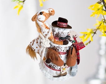 May include: A diaper cake decorated with a stuffed horse, cowboy hat, boots, and bandana. The cake is made of diapers and is decorated with a white lace trim.
