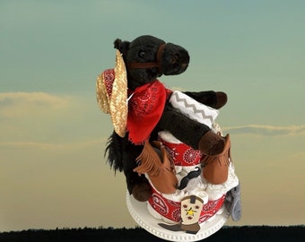 May include: A black horse soft toy wearing a red bandana and a straw cowboy hat sits atop a nappy cake decorated with red and white bandanas, brown booties, and a moustache.