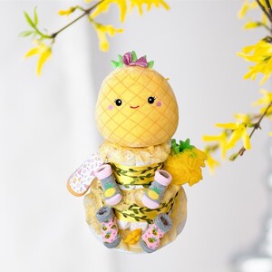 May include: A yellow pineapple-shaped plush toy with a pink bow sits atop a diaper cake decorated with yellow and green ribbon, white lace, and baby socks with pineapple designs.