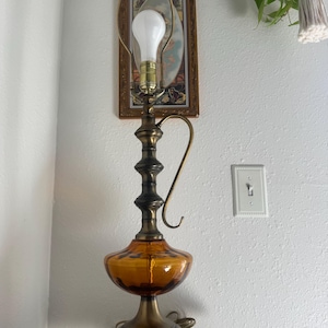 May include: A table lamp featuring a brass-toned metal base and a central handle, topped with a harp and a single white light bulb. The lower section consists of a translucent amber-colored glass body with a rounded, wide shape and a circular base.