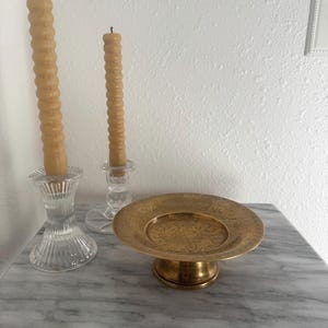 May include: A still life arrangement featuring two tall, textured, light brown candles in clear glass candle holders. A small, round, gold-colored decorative plate with an embossed floral design sits on a marble surface.