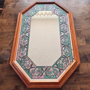 May include: An octagonal wall mirror featuring a light brown wooden frame. The mirror glass is bordered by a decorative stained glass style pattern in shades of teal, pink, and white, depicting repeating floral and leaf motifs.