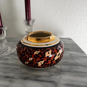 May include: A round ceramic vase featuring a bold leopard print pattern in shades of brown, black, and cream. The vase has a wide, smooth gold-toned metallic rim at the top opening, providing a distinct contrast to the animal print body below.
