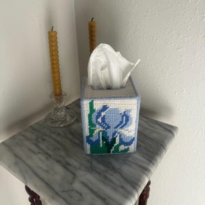 May include: A rectangular tissue box cover crafted from white plastic canvas with a needlepoint design featuring a blue iris flower with green leaves and stems. The cover has a light blue border and holds a stack of white facial tissues.