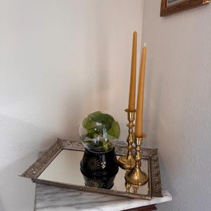 Puede incluir: Una bandeja rectangular con espejo y marco metálico ornamentado sostiene una maceta negra con un diseño de sol y estrellas doradas que contiene una planta verde bajo una cúpula de cristal, junto a dos velas amarillas en candelabros de latón.