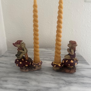 May include: Two whimsical candle holders with twisted yellow candles. Each holder features a fairy figurine seated on a mushroom with brown and white spots. The candle holders are on a marble surface.