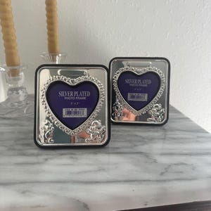 Może przedstawiać: Dwie posrebrzane ramki na zdjęcia z otworami w kształcie serca i dekoracyjnymi akcentami kwiatowymi i cherubinami. Każda ramka ma wymiary 7,6 cm x 7,6 cm i czarną obwódkę. Ramki są wystawione na stole z marmurowym blatem.