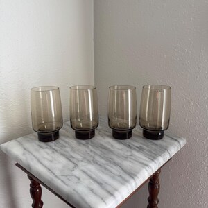 May include: A set of four matching drinking glasses made of translucent smoke-coloured glass. Each glass features a cylindrical body that tapers slightly towards the rim and sits on a thick, weighted base with a darker, solid brown glass bottom.