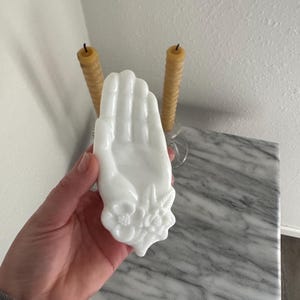 Op de afbeelding: Een wit, handvormig decoratief object met bloemendetails aan de basis. Twee gele kaarsen staan op de achtergrond. Het object staat op een marmeren oppervlak.