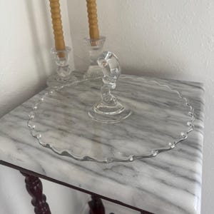 Op de afbeelding: Een heldere glazen taartstandaard met een geschulpte rand, rustend op een tafel met marmeren blad. Twee glazen kandelaars met brandende, bruine kaarsen staan op de achtergrond. De tafel heeft donkerbruine poten.