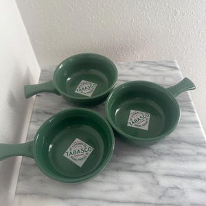 Puede incluir: Tres cuencos de cerámica verdes con asas, cada uno con una pegatina del logotipo de la marca "TABASCO" en blanco y verde. Los cuencos están colocados sobre una superficie de mármol blanco y gris.