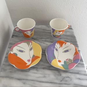 May include: A pair of teacups and saucers featuring a colourful abstract design of a woman's face. The saucers have yellow, orange, and purple backgrounds. The cups have pink, yellow, and purple backgrounds. The set is on a marble surface.