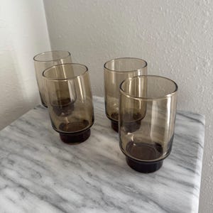 May include: A set of four cylindrical drinking glasses made of translucent smoke-coloured glass. Each glass features a slightly thicker, darker base that transitions into a lighter, transparent body, creating a uniform appearance across all four items.