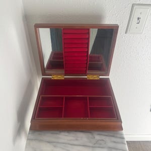 May include: Open wooden jewelry box with a red velvet interior and a mirrored lid. The box sits on a marble-topped table with a dark wood base. The box is open, revealing compartments for storage.