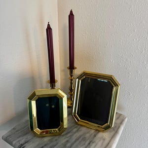 May include: Two polished gold-tone metal picture frames with octagonal borders and dark inserts, accompanied by two tall, slender, ribbed burgundy taper candles, one of which is held in a matching gold-tone metal candlestick holder.