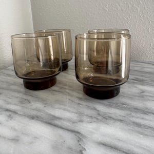 May include: A set of four short drinking glasses made of translucent brown glass. Each glass features a cylindrical body with a slightly wider, solid brown base that provides stability. The glass has a smooth, uniform finish throughout.