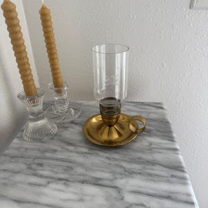 May include: A marble-topped table displays two clear glass candle holders with spiral-shaped beeswax candles. A brass candle holder with a glass hurricane lamp is also on the table. The table has a grey and white veined pattern.
