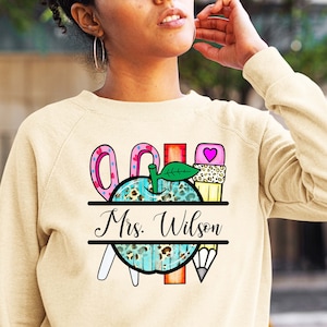 Puede incluir: Sudadera beige con un diseño colorido con temática de profesor. El diseño incluye tijeras, una manzana, una magdalena y un lápiz. El nombre "Mrs. Wilson" está impreso debajo.