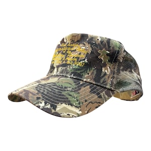 Op de afbeelding: Een camouflage baseballpet met een mesh achterkant en een gebogen klep. De voorkant is voorzien van geel geborduurde tekst met de tekst 21st Annual International Order of the Alhambra Festival & Parade 1st 2nd & 3rd 2007 in een cursief lettertype.