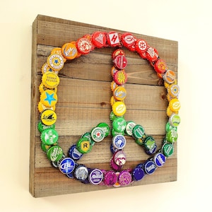 May include: A peace sign made from colourful bottle caps on a wooden background. The caps are arranged in a rainbow pattern, starting with red at the top and ending with purple at the bottom.