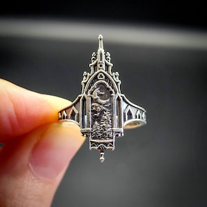 May include: A silver ring with a detailed design of a gothic cathedral with a crescent moon and stars in the window. The ring is worn on a finger.
