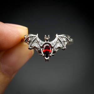 Può includere: Un anello d'argento a forma di pipistrello con una pietra preziosa rossa al centro. Le ali del pipistrello sono dettagliate con un design testurizzato.