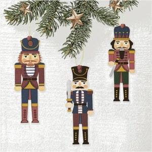 May include: Three wooden Christmas tree decorations shaped as nutcrackers, suspended from a pine branch with gold star ornaments. The nutcrackers are in red, blue, and gold, each with a unique uniform and facial expression. A festive holiday decoration.