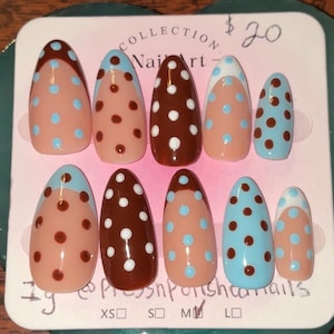 May include: A set of ten almond-shaped press-on nails displayed on a white card. The nails feature a mix of nude, brown, and light blue base colors with contrasting polka dot patterns and French-style tips. The card includes text and size options.