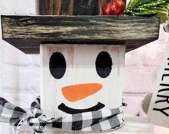 Puede incluir: Una decoración de muñeco de nieve de madera con una bufanda a cuadros en blanco y negro, ojos negros, una nariz naranja y tres botones negros. El muñeco de nieve está de pie sobre una base de madera blanca.