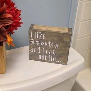 May include: A wooden block sign with the text "I like big butts and I can not lie..." sits on a white toilet. A vase of fall-colored flowers is to the left of the sign. The flowers include orange, red, and brown.