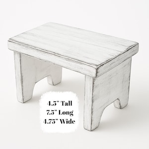 May include: A small, white, distressed wooden step stool. The stool is 4.5 inches tall, 7.5 inches long, and 4.75 inches wide. It has a rectangular top and four sturdy legs. The white paint is slightly worn, giving it a rustic appearance.