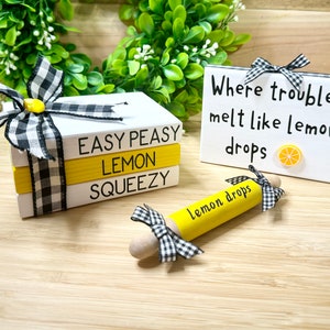 Op de afbeelding: Een set van drie gestapelde houten blokken met de woorden "Easy Peasy Lemon Squeezy" erop geschreven. De blokken zijn versierd met een zwart-wit geruit lint. Een klein houten bord met de tekst "Where troubles melt like lemon drops" en een citroenschijfje afbeelding is ook afgebeeld. Een gele deegroller met de tekst "lemon drops" is ook afgebeeld.