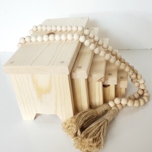 May include: A set of four unfinished wooden boxes with rounded edges, stacked on top of each other. The boxes are decorated with a strand of wooden beads and two natural fiber tassels.