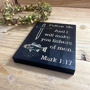 K&ouml;nnte beinhalten: Schwarze Holzplakette mit graviertem Text und Fischillustration. Der Text lautet: &bdquo;Folge mir, und ich will euch zu Menschenfischern machen. Markus 1:17.&ldquo; Ein Kreuz befindet sich &uuml;ber dem Text. Im Hintergrund befindet sich ein dekorativer Kranz.